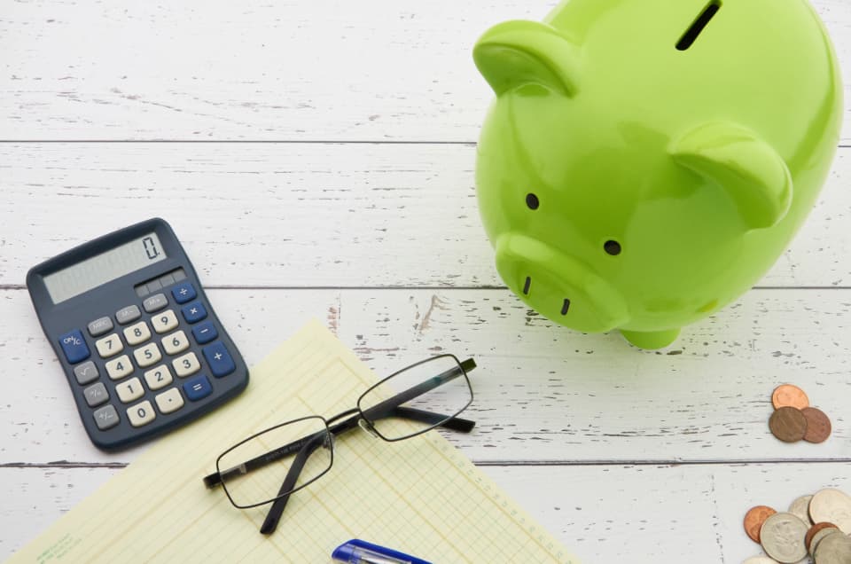 Green piggy bank next to a calculator, reading glasses, a ledger pad and loose coins